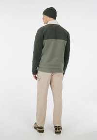 Groene fleece pullover met een lichtgroen bovenstuk, in combinatie met beige cargo broeken en groene wandelschoenen. Model draagt een bijpassende beanie.
