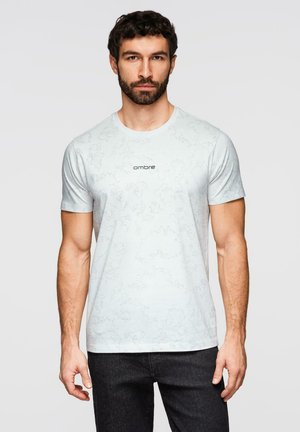 Man met baard draagt een lichtgrijs T-shirt met korte mouwen, subtiele abstracte patronen en de tekst "ombre", gecombineerd met een donkere spijkerbroek.