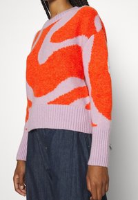Pull en tissu duveteux avec un motif audacieux orange et lavande, col rond et poignets côtelés. La texture douce complète le design vibrant.