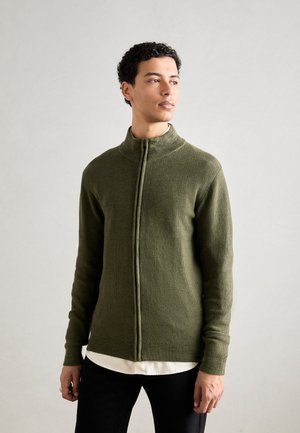 Maslinasto zelena zip-up dukserica, teksturirani pletenac, visoki ovratnik, dugi rukavi, prilagođen dizajn. Nosena preko bijele košulje i crnih hlača.