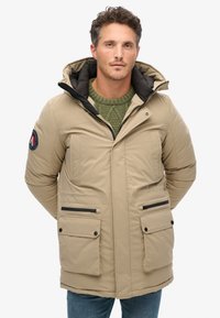 Parka beige con capucha, dos bolsillos frontales, detalles en negro en la cremallera y suéter verde de punto debajo. Textura suave y diseño acolchado.