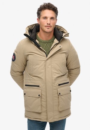 Parka beige con capucha, dos bolsillos frontales, detalles en negro en la cremallera y suéter verde de punto debajo. Textura suave y diseño acolchado.