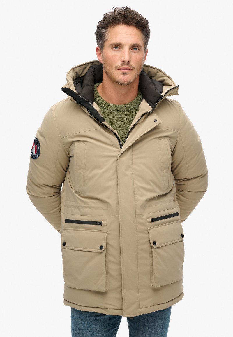 Parka beige con capucha, dos bolsillos frontales, detalles en negro en la cremallera y suéter verde de punto debajo. Textura suave y diseño acolchado.