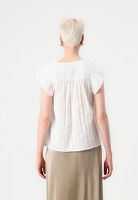 Blusa bianca a maniche corte con spalle arricciate, caratterizzata da righe verticali e una parte posteriore arricciata. Abbinata a una gonna beige plissettata.