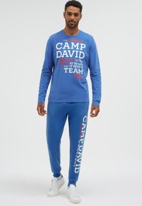 Mann trägt blaues Camp David Sweatshirt und passende Jogginghose mit weißen Turnschuhen, steht vor einfarbigem Hintergrund.