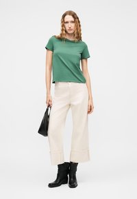 Γυναίκα με κυματιστά μαλλιά, φορώντας πράσινο κοντομάνικο τοπ, φαρδιά κρεμ παντελόνια, μαύρες μποτάκια αστραγάλου, κρατώντας μια μικρή μαύρη τσάντα χειρός.