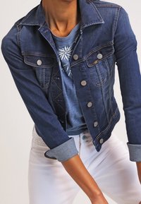 Tummansininen farkkutakki, jossa on istuva muotoilu, klassinen kaulus ja napitus. Päällä vaaleansininen paita ja valkoiset housut.