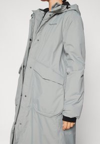 Manteau imperméable gris clair avec capuche, doté de boutons-pression, de grandes poches avant et de poignets élastiques pour une fonctionnalité accrue.