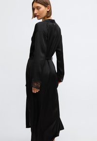 Čierny saténový župan s dlhými rukávmi, čipkovými manžetami, opaskom v páse a vysokým rázporkom. Má okrúhly golier a gombík na zadnej strane.