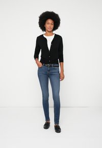 Moteris su natūraliu afro šukuosena stovi prieš baltą foną, vilkinti juodą kardiganą, baltą marškinėlį, mėlynus džinsus, juodą diržą ir juodas loafer batus.