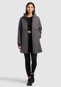 Ženska, ki nosi sivo vetrovko s kapuco do sredine stegen, črno kratko majico, črne legice in črne športne copate, stoji pred sivim ozadjem.