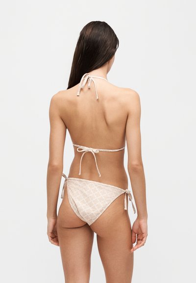 Femme aux longs cheveux foncés portant un bikini beige clair à motifs, noué au cou, dans le dos et aux hanches, debout, face à dos sur fond uni.