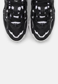 Ett par svarta och vita sportskor med detaljer i nät och läder, sedda uppifrån mot en enkel ljusgrå bakgrund.