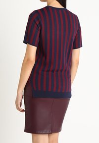 Une femme portant un haut à rayures verticales bleu marine et bordeaux à manches courtes, associé à une jupe en cuir bordeaux de longueur genou, se tenant devant un fond blanc.