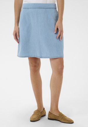 Femme portant une jupe en jean bleu clair jusqu'aux genoux et des mocassins en daim beige, debout sur un fond blanc uni.