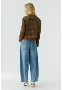 Vrouw staande met de rug naar de camera, gekleed in een bruine geruite jas, losse lichtblauwe jeans en witte hoge hakken op een effen achtergrond.