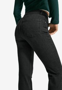 Frau in schwarzen, enganliegenden Jeans und einem dunkelgrünen, cropped Langarmshirt, die vor einem weißen Hintergrund steht.