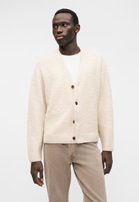 Πλεκτή ζακέτα σε κρεμ απόχρωση με υφή, με λαιμόκοψη σε σχήμα V, μακριά μανίκια και τρία ξύλινα κουμπιά στο μπροστινό μέρος.