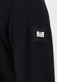 Zwarte sweatshirt met een gestructureerde stof, geribde manchetten en een vierkante label met "WEEKEND OFFENDER" op de mouw.