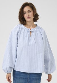 Blouse rayée bleu clair et blanche avec des manches bouffantes, un col froncé et une ouverture en forme de clé à l'avant. Tissu en coton, coupe décontractée.