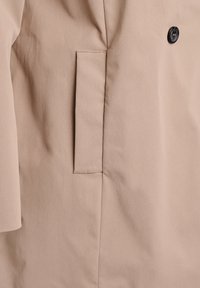 Beżowa kieszeń płaszcza z pionową, prostokątną patką i czarnym guzikiem na gładkiej tkaninie.