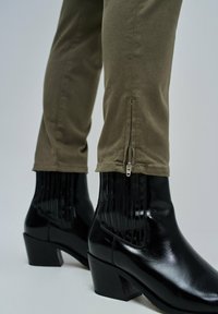 Bottines à talons noirs avec une texture brillante et des accents rayés verticaux, associées à un pantalon vert olive avec un détail de fermeture éclair sur le côté.