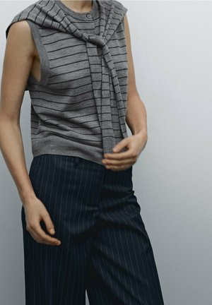 Person wearing a sleeveless gray striped sweater with a matching sweater tied around shoulders and navy pinstriped pants.