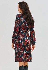 Květinové šaty z tmavé látky s červenými, oranžovými a bílými vzory; dlouhé rukávy; úzký pas; do délky ke kolenům; spadá elegantně přes boky.