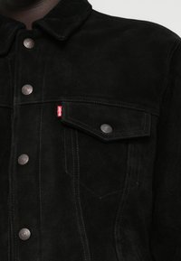 Μαύρο σουέντ μπουφάν με αιχμηρό γιακά, μπροστινές τσέπες και μεταλλικά κουμπιά με κούμπωμα. Διαθέτει αντίθετη ραφή και μια ετικέτα με το λογότυπο σε κόκκινο χρώμα.