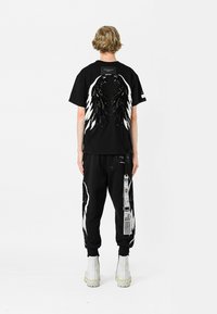 Camiseta negra con gráficos de alas blancas y detalles de lentejuelas en la parte de atrás, combinada con pantalones negros que presentan patrones de alas similares y un diseño de etiqueta.