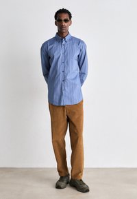 Camicia azzurro chiaro a righe con maniche lunghe, abbinata a pantaloni in velluto a coste marroni. Il modello indossa occhiali da sole scuri e sneaker verdi.