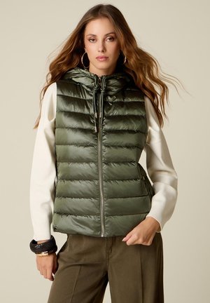 Donna che indossa un gilet imbottito verde oliva senza maniche con cappuccio sopra una maglia crema a maniche lunghe, pantaloni marroni e grandi orecchini a cerchio.