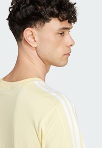 Camiseta de algodón de un suave amarillo con diseño de tres rayas blancas en los hombros. Cuello redondo y un corte relajado, presentada de lado.
