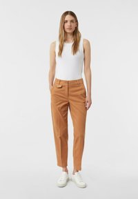 Donna in piedi che indossa un top bianco senza maniche, pantaloni color cammello con tasche e sneakers bianche, su uno sfondo bianco uniforme.