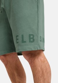Grüne Baumwollshorts mit weicher Textur, versehen mit dunkelgrüner Schrift "ELB" am linken Bein und "SNA" am Saum, knielange Ausführung.