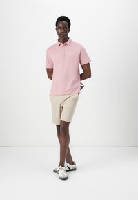 Light pink polo shirt with a collar, short sleeves, and embroidered logo. Beige shorts and gray sneakers complete the outfit.