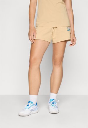 Beige shorts laget av mykt stoff, med en brodert logo. Brukt med hvite sneakers pyntet med blå detaljer og hvite ankelstrømper.