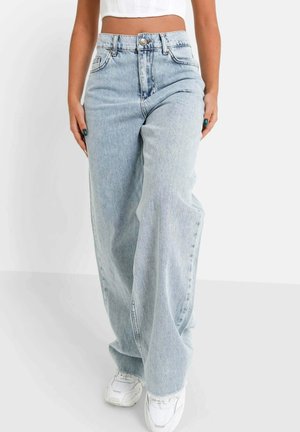 Person trägt hellblaue Jeans mit weitem Bein, weiße Turnschuhe und ein weißes, kurzes Oberteil und steht vor einem neutralen Hintergrund.