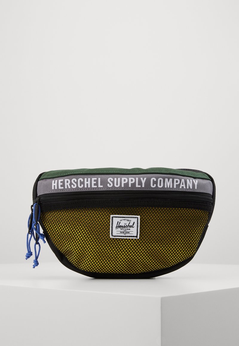 Klein ritstasje met groene, grijze en gele netpanelen, blauwe ritstrekkers en "Herschel Supply Company" branding.