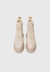 Béžové semišové boty na šněrování s tlustou gumovou podrážkou a kožešinovou podšívkou, zobrazené shora na bílém pozadí.