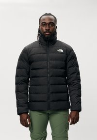 Chaqueta de plumas negra con cuello alto, cierre de cremallera y diseño acolchado. Logo en el lado izquierdo del pecho. Combinada con pantalones cargo verdes.
