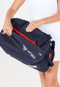 Hænder der åbner en sort og rød sportstaske, der holdes af en person iført et hvidt outfit mod en ensfarvet baggrund.