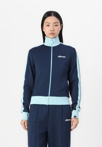 Mornarska trening jakna z svetlo modrimi detajli, visokim ovratnikom, z zadrgo spredaj in rebrastimi manšetami. Vidno logotip na prsih. Mehka, športna tkanina.