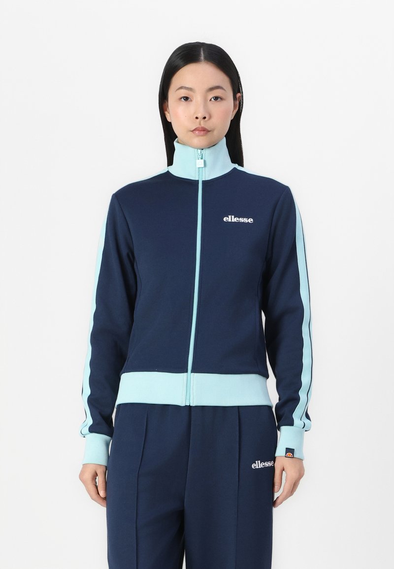 Mornarska trening jakna z svetlo modrimi detajli, visokim ovratnikom, z zadrgo spredaj in rebrastimi manšetami. Vidno logotip na prsih. Mehka, športna tkanina.