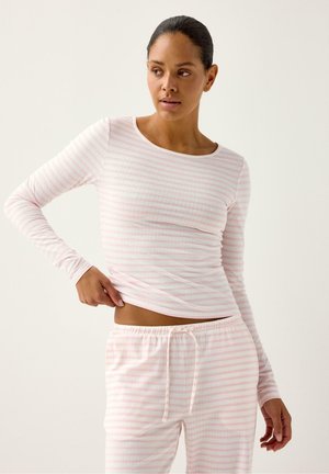 Kvinde justerer lyserød og hvidstribet langærmet top og matchende bukser med snor foran en ensartet lys baggrund.