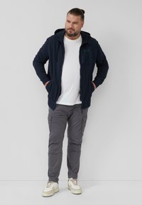 Marineblauwe zip-up hoodie met een voorkantzak, gecombineerd met grijze cargobroeken en witte sneakers, voorzien van een relaxte pasvorm en een verstelbare capuchon.