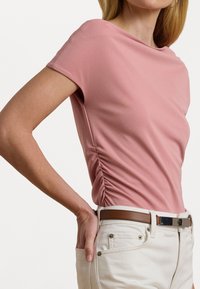 T-shirt rosa a coste con maniche corte, dettaglio arricciato sui lati e scollatura a barca. Abbinato a pantaloni bianchi e una cintura marrone.
