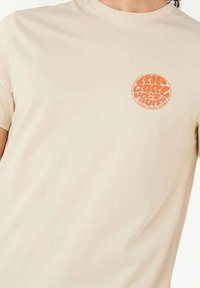 Beige bomuld t-shirt med korte ærmer, der har et cirkulært orange grafisk logo på venstre brystområde. Glat tekstur, standard pasform.