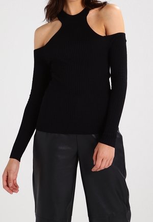 Frau in einem schwarzen, gerippten Pullover mit kalten Schultern und langen Ärmeln, kombiniert mit schwarzen Leder-Shorts, vor einem schlichten Hintergrund.