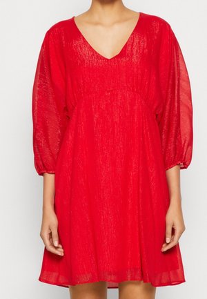 Kobieta w luźnej, błyszczącej czerwonej sukience z dekoltem w serek i trójczynowymi, przezroczystymi rękawami, stojąca na jednolitym białym tle.